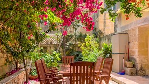 A patio with wooden chairs and a table, surrounded by greenery and a stone wall. - Bougainvillea Breeze & Marina View (Cospicua)