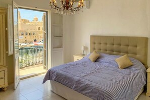 A bedroom with a large bed, a chandelier, a nightstand, and a view of a cityscape through an open door.