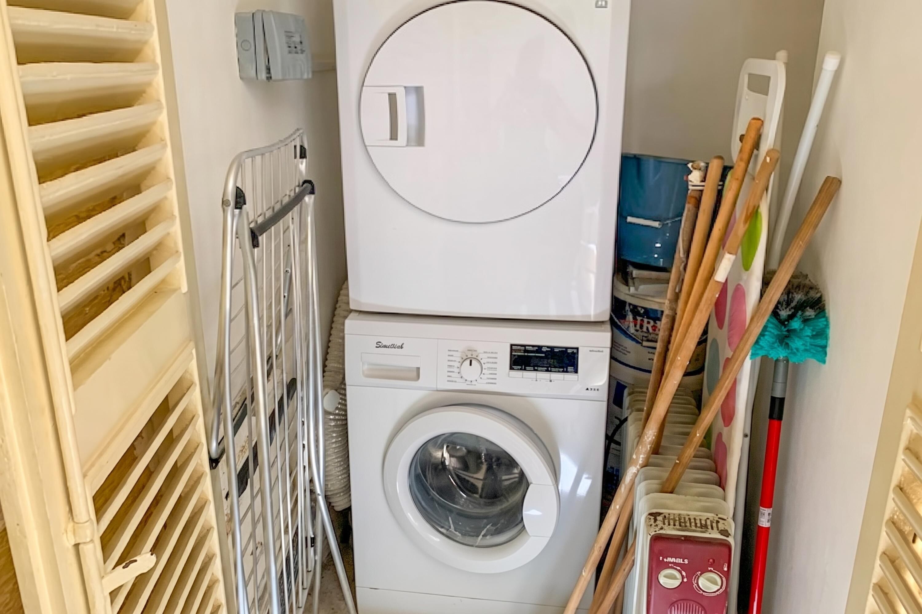 Uma lavanderia compacta com máquina de lavar e secadora frontais, um varal de metal e um esfregão encostado na parede.