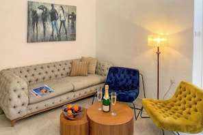 A living room with a tufted sofa, a blue armchair, a yellow armchair, a small wooden table with a bowl of fruit and a bottle of champagne, and a floor lamp.