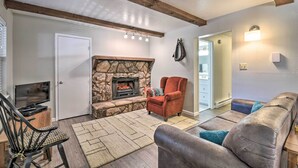 A living room with a stone fireplace, a red armchair, a grey sofa, and a wooden ceiling beam. - Angel Fire Condo w/ Fireplace < 1 Mi to Ski Resort (Angel Fire)