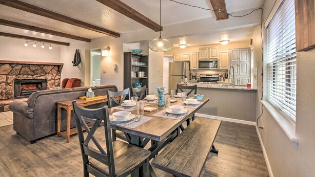 A dining area with a long wooden table set for four, surrounded by dark wooden chairs. A stone fireplace is visible in the background, and the room features exposed wooden beams on the ceiling. - Angel Fire Condo w/ Fireplace < 1 Mi to Ski Resort (Angel Fire)