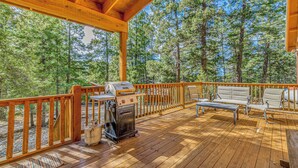 Una terraza de madera con una parrilla, asientos al aire libre y vistas a un bosque.