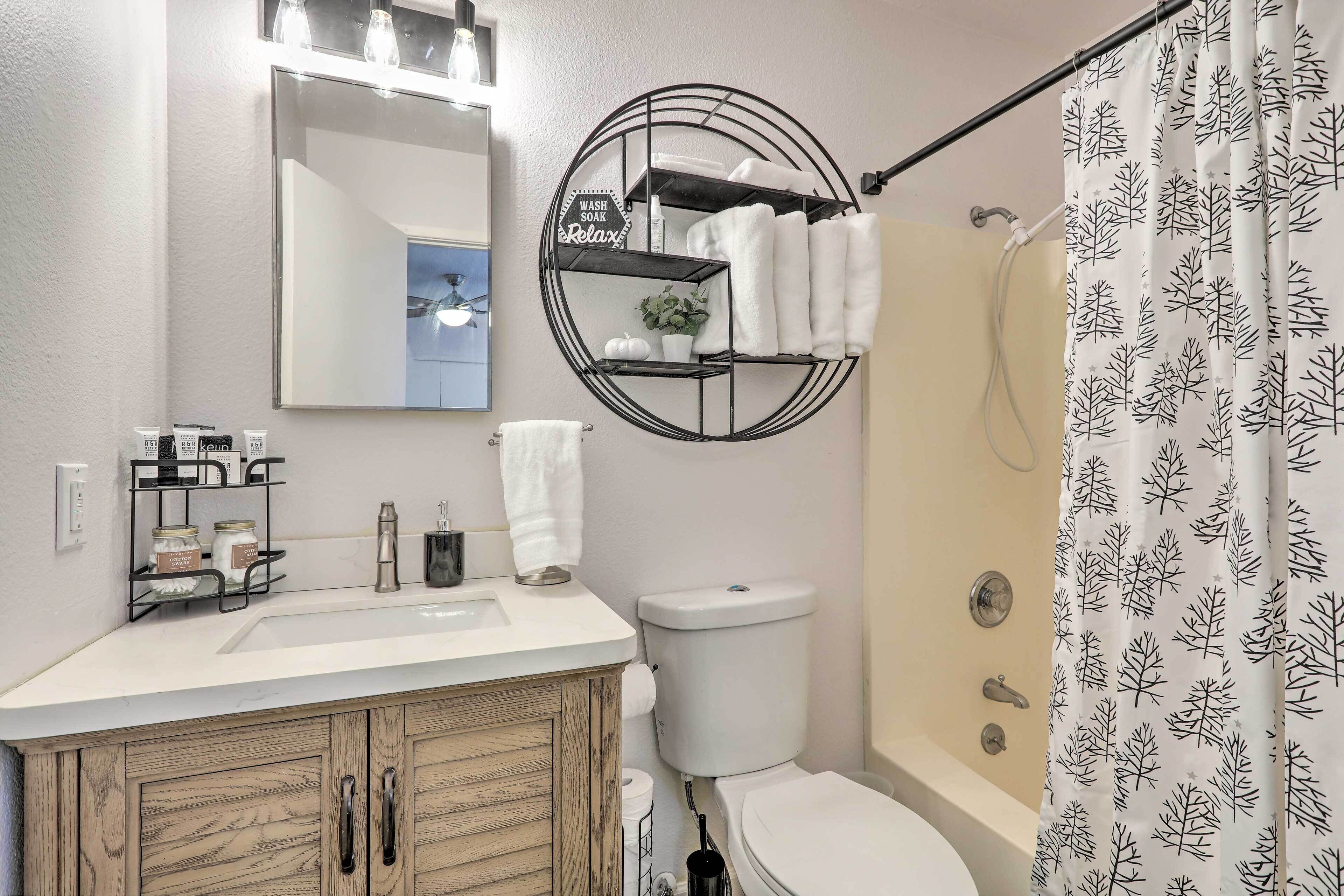A bathroom with a white sink, wooden vanity, a toilet, a shower curtain with a leaf pattern, and a shelf with towels and a sign.
