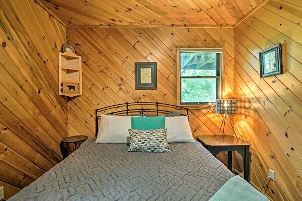 A cozy bedroom with a wooden ceiling, a bed with a quilted blanket, two pillows, a small bedside table with a lamp, and a window with a view of greenery.