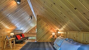 An attic bedroom with a sloped ceiling, wooden walls, a bed with blue bedding, a sofa with red cushions, and two bedside lamps. - Near Snowmobiling! Riverfront Traverse City Cabin (Traverse City)