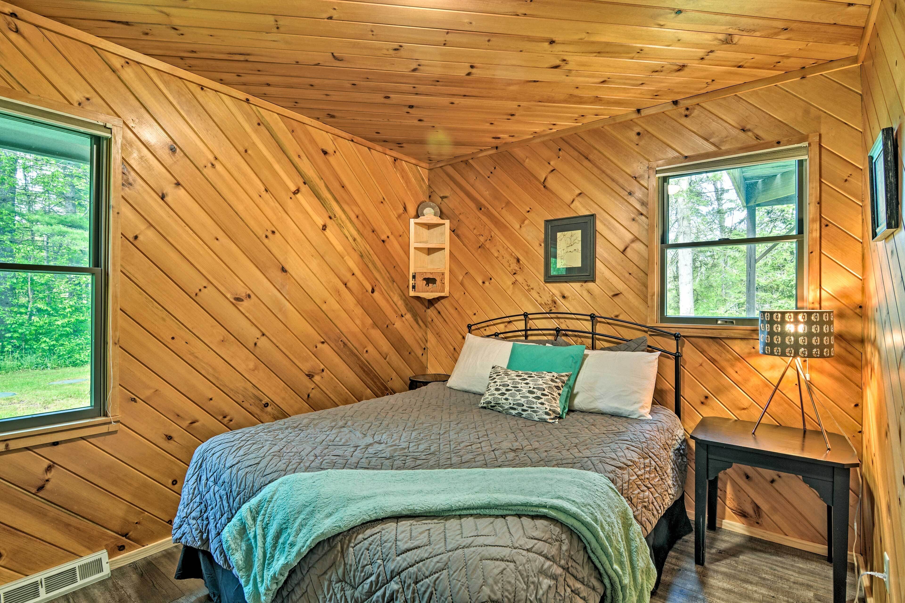 Une chambre dans un chalet en bois, avec un lit, une table de chevet, une étagère fixée au mur et deux fenêtres.