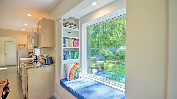 Una cocina con armarios empotrados, un rincón ventana con cojín de arcoíris y vistas a un paisaje verde.