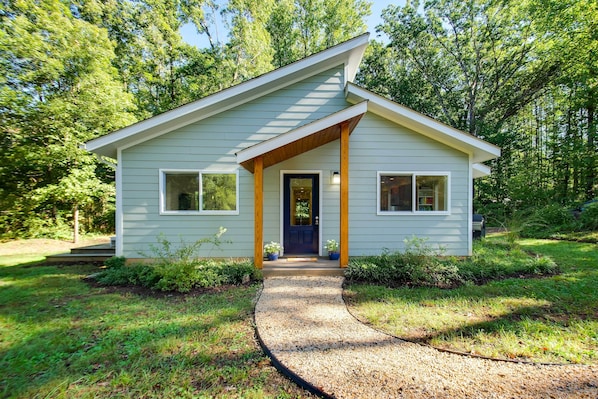 Une petite maison de plain-pied, dotée d’un toit en pente, d’un porche à l’avant avec des poutres en bois et d’un chemin de gravier menant à l’entrée.