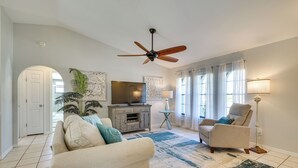Un salon avec un ventilateur de plafond, un téléviseur, un canapé, un fauteuil, une lampe et un tapis.