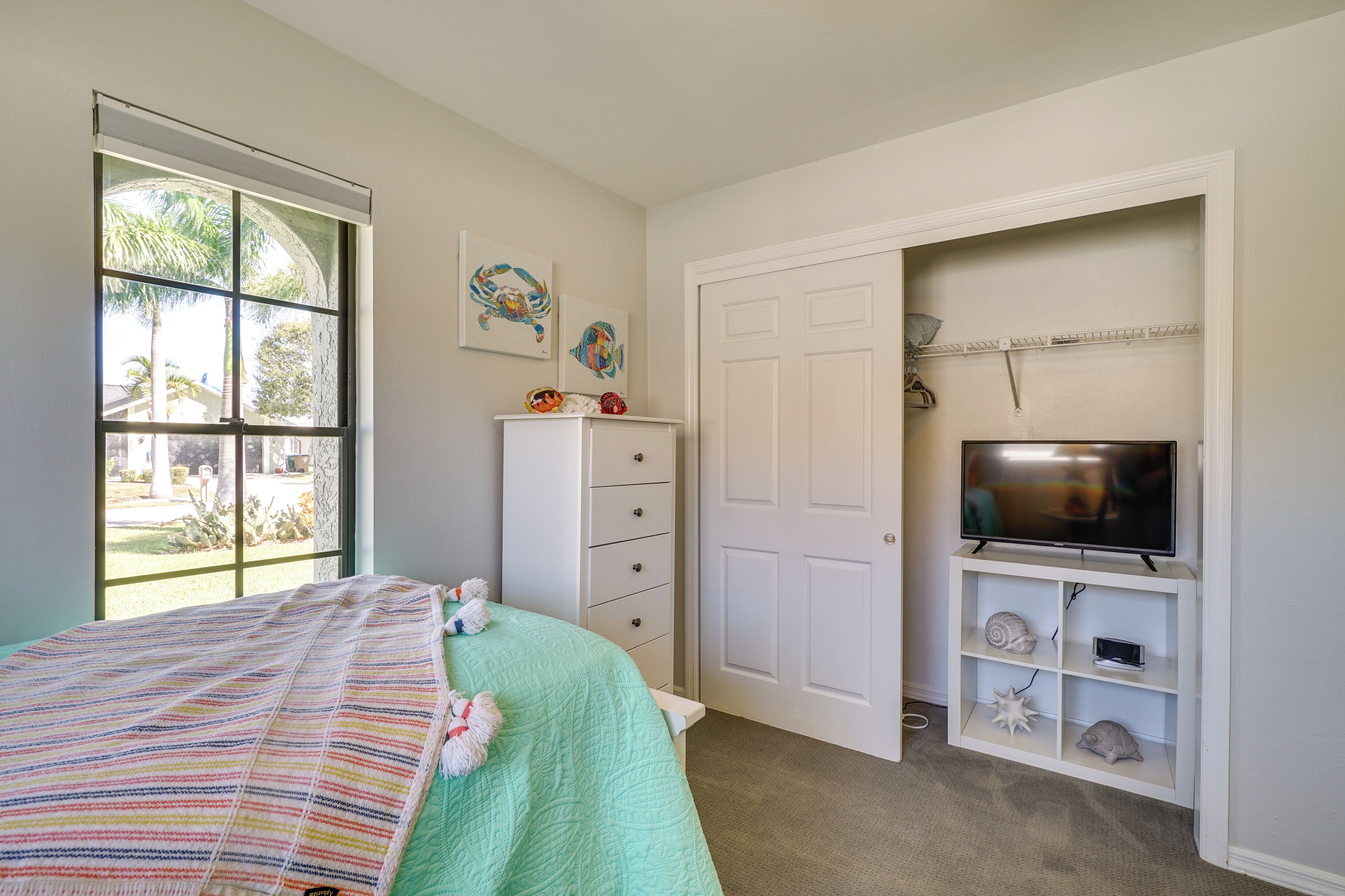 Une chambre à coucher avec un lit, une commode, un téléviseur posé sur une étagère et une fenêtre donnant sur des palmiers.