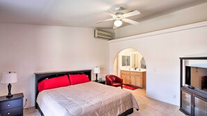 A bedroom with a bed, a nightstand, a lamp, a ceiling fan, an air conditioning unit, a red chair, a TV, and a bathroom visible through an arched doorway.