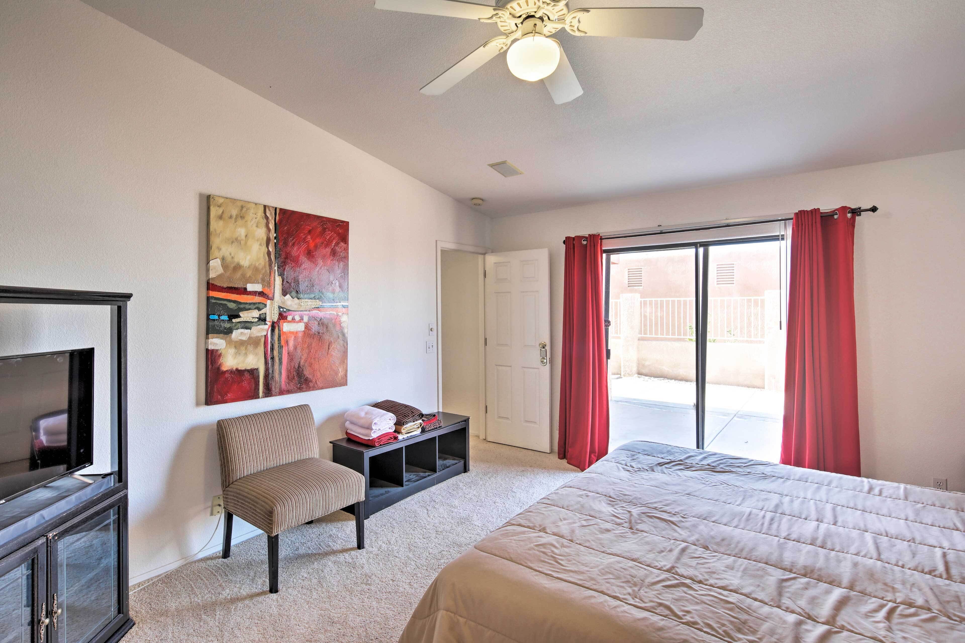 A bedroom with a bed, a chair, a TV, a ceiling fan, a door, and a window with red curtains.