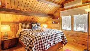 Une chambre confortable dans un chalet en bois, avec une couverture à carreaux sur le lit, une table de chevet avec une lampe et une fenêtre donnant sur les arbres.