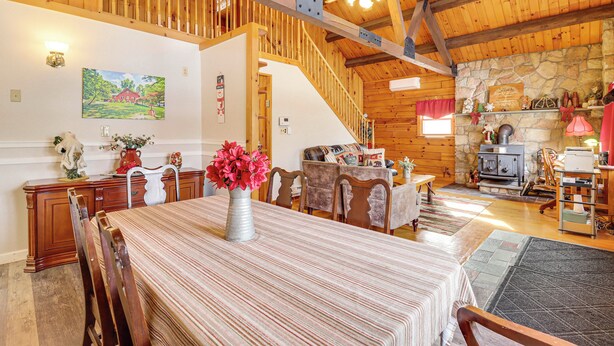 Un vaste espace dédié à la salle à manger, avec une longue table, des chaises en bois et un vase rempli de fleurs. Un escalier mène à l’étage supérieur, et une cheminée en pierre est visible au fond.