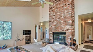 A spacious living room with a brick fireplace, a ceiling fan, and a large window. - Pine Mountain Club Cottage W/wraparound Deck! (Pine Mountain Club)