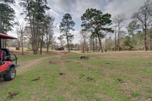 A rural landscape with a dirt path leading to a wooden structure, surrounded by tall trees and open grassy areas.