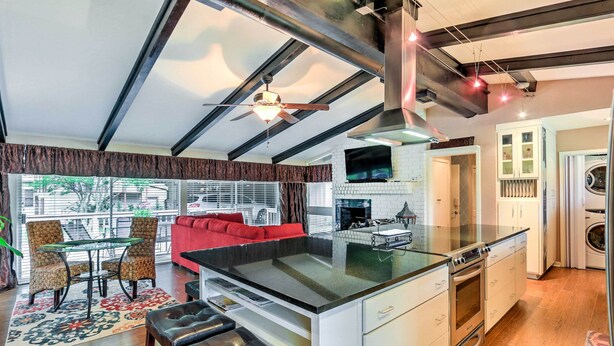 Een moderne keuken met een centraal eiland, een eethoek met een glazen tafel en stoelen, een rode bank en een groot raam met jaloezieën.