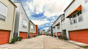 Uma fileira de edifícios em estilo industrial, com grandes portões de garagem, céu claro e um caminho central.