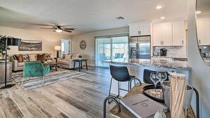 A modern living room with a sofa, coffee table, and chairs, a dining area with a table and chairs, and a kitchen with white cabinets and a refrigerator.