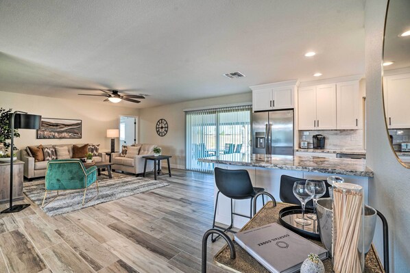 A modern living room with a sofa, coffee table, and chairs, a dining area with a table and chairs, and a kitchen with white cabinets and a refrigerator.