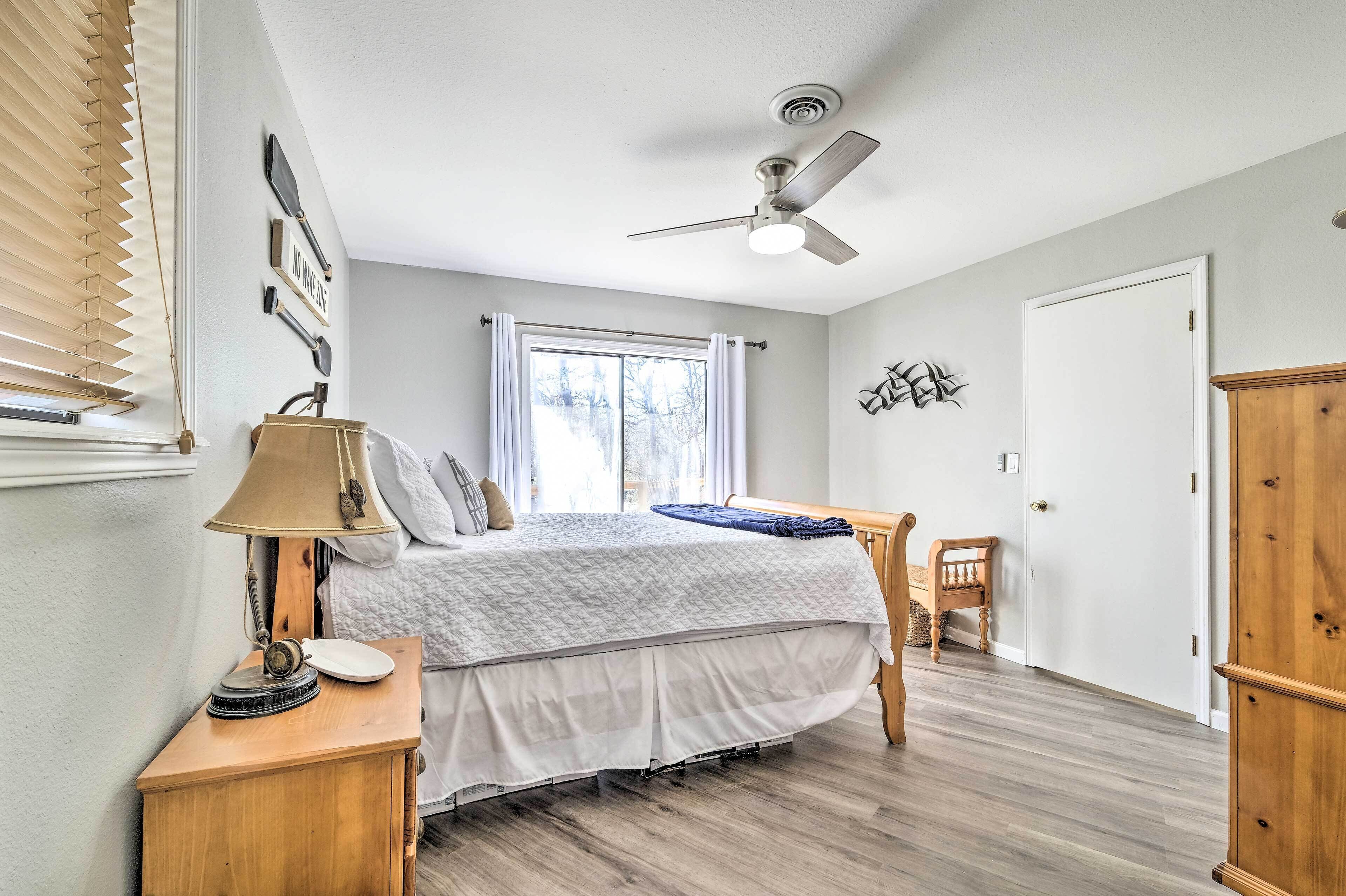 A bedroom with a bed, bedside table, lamp, window with blinds, ceiling fan, and a door.
