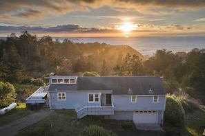A two-story house with a dock, surrounded by forested hills and a view of the ocean at sunset. - Hot Tub + Ocean Views: Spacious Irish Beach Home! (Manchester)