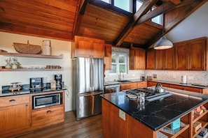 A modern kitchen with wooden cabinets, a stainless steel refrigerator, a built-in oven, and a large island with a black countertop. - Hot Tub + Ocean Views: Spacious Irish Beach Home! (Manchester)