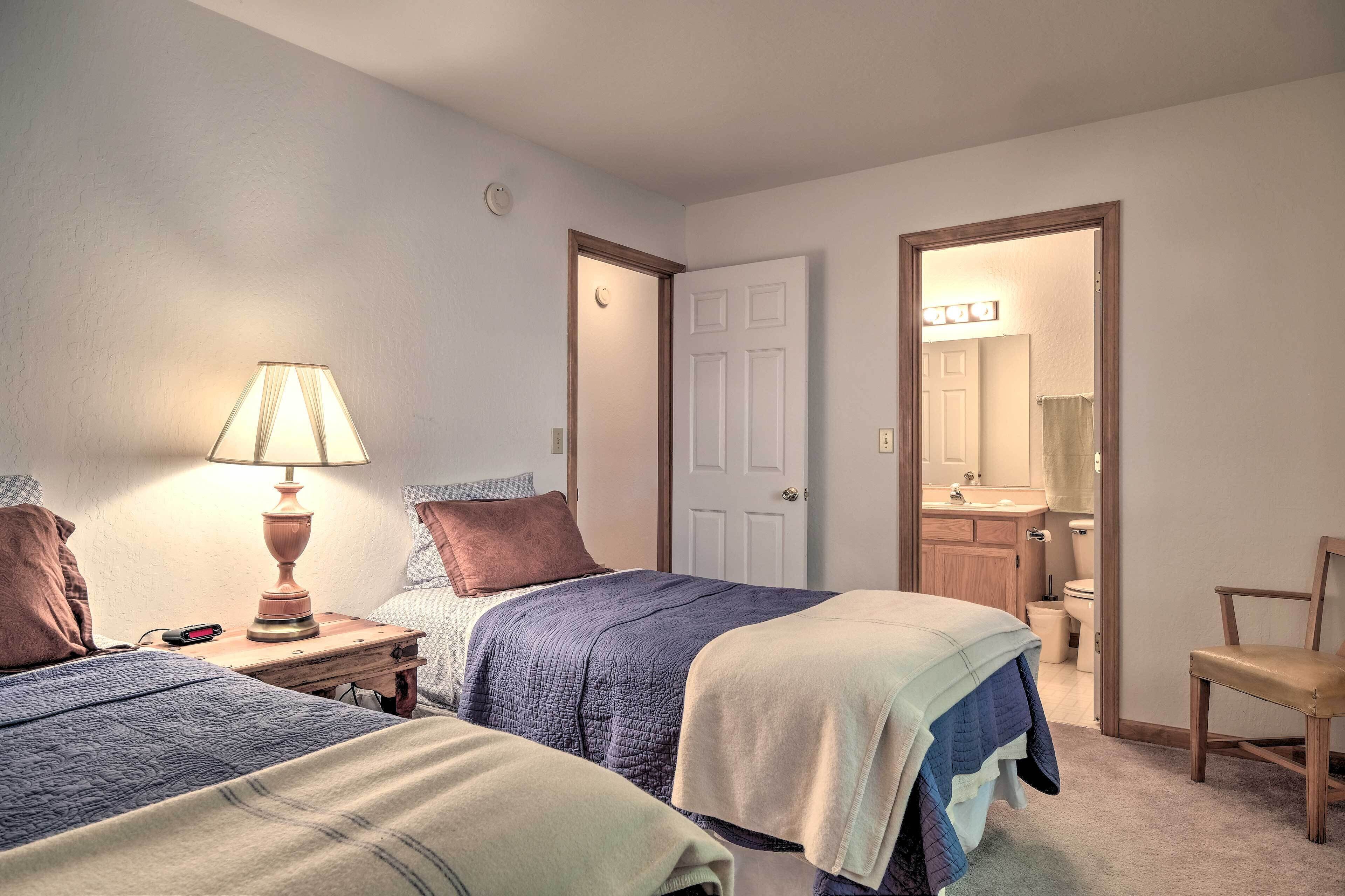 A bedroom with two beds, a nightstand with a lamp, a chair, and a bathroom visible through an open door.