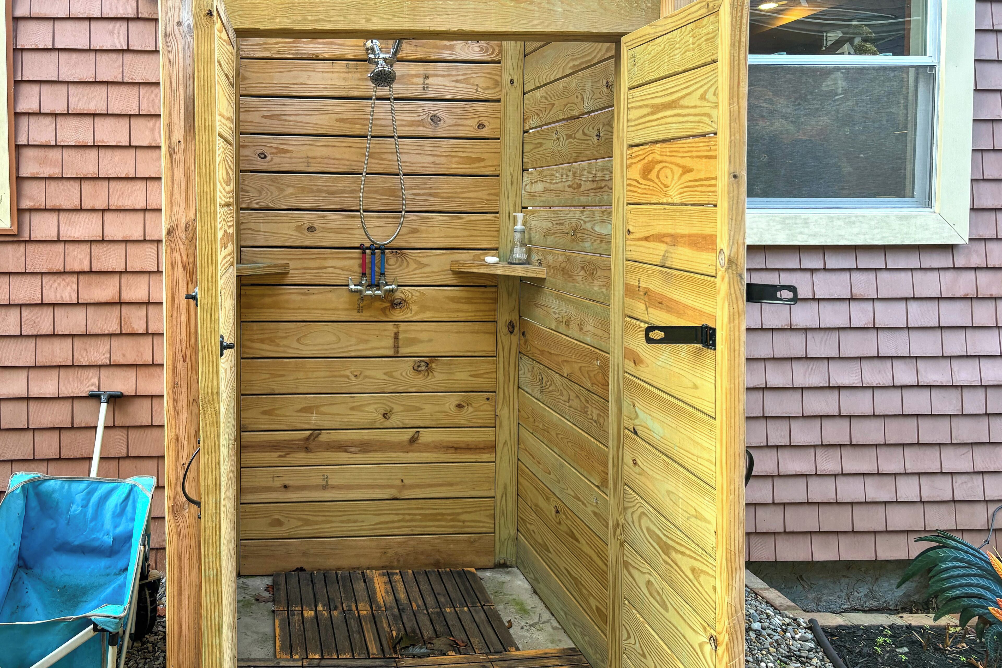 Une cabine de douche en bois avec une fenêtre, une petite étagère avec un distributeur de savon, et une brouette bleue à l’extérieur.