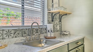 A kitchen with a sink, a window with blinds, a shelf with glassware, and a patterned backsplash. - El Paso Cottage - Walk to UMC & Texas Tech! (El Paso)
