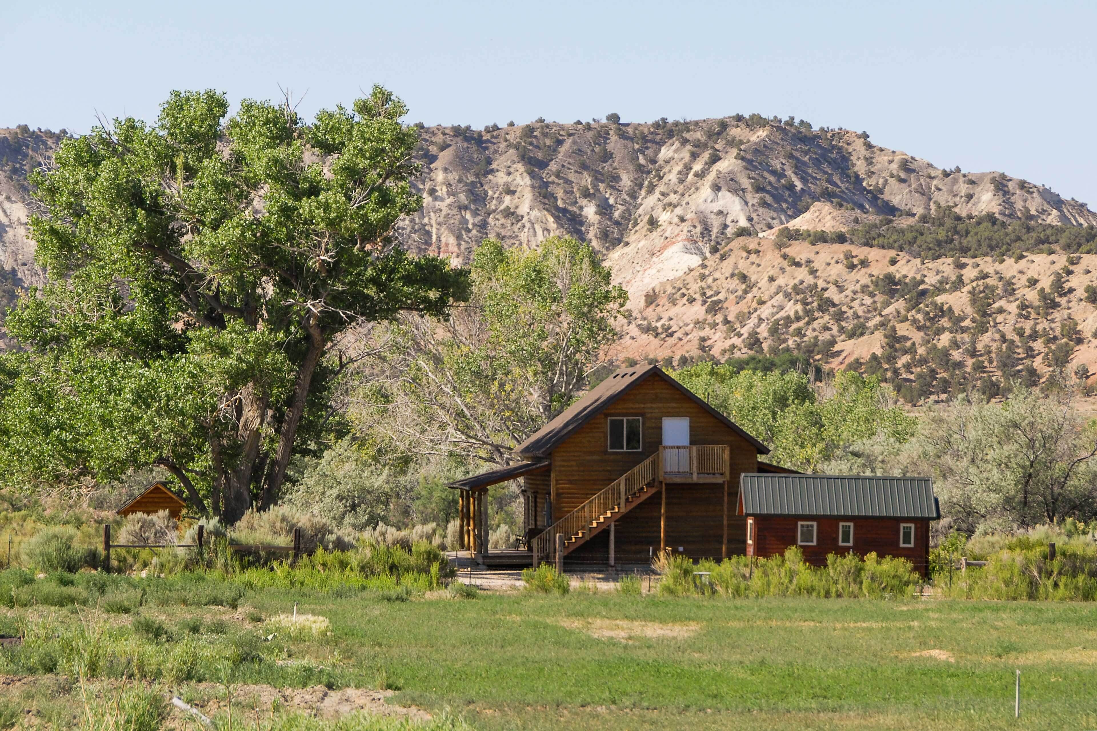 A wooden cabin with a porch and staircase, surrounded by greenery and a mountainous backdrop.