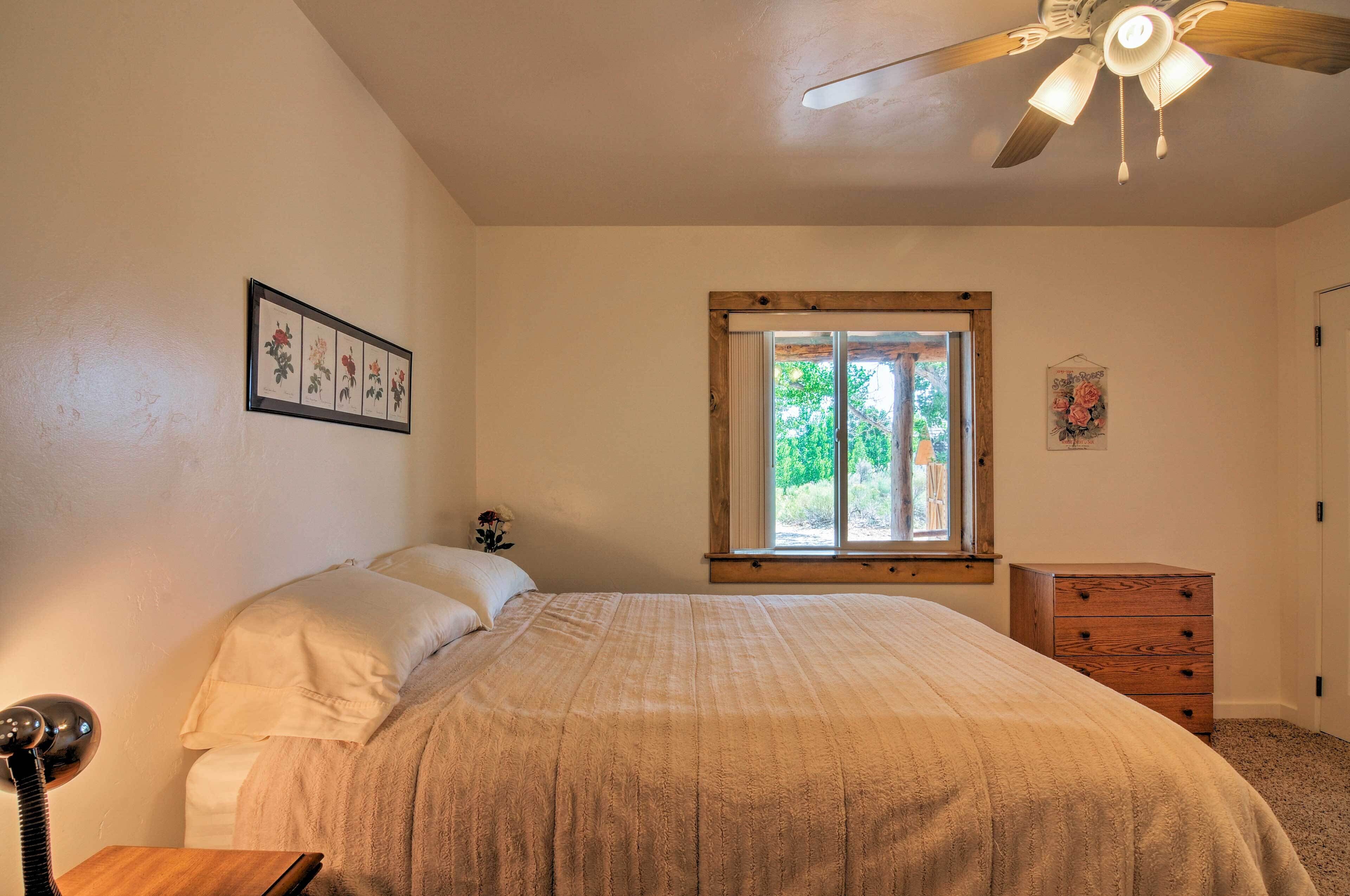 Un dormitorio con cama, cómoda de madera, ventilador de techo, ventana con vistas a follaje, y obras de arte enmarcadas en la pared.