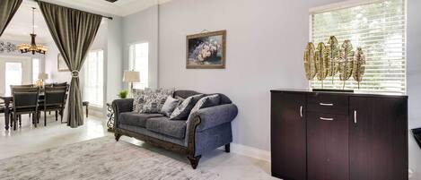 A spacious living room with a grey sofa, dark wooden cabinet, and a dining area with a chandelier.