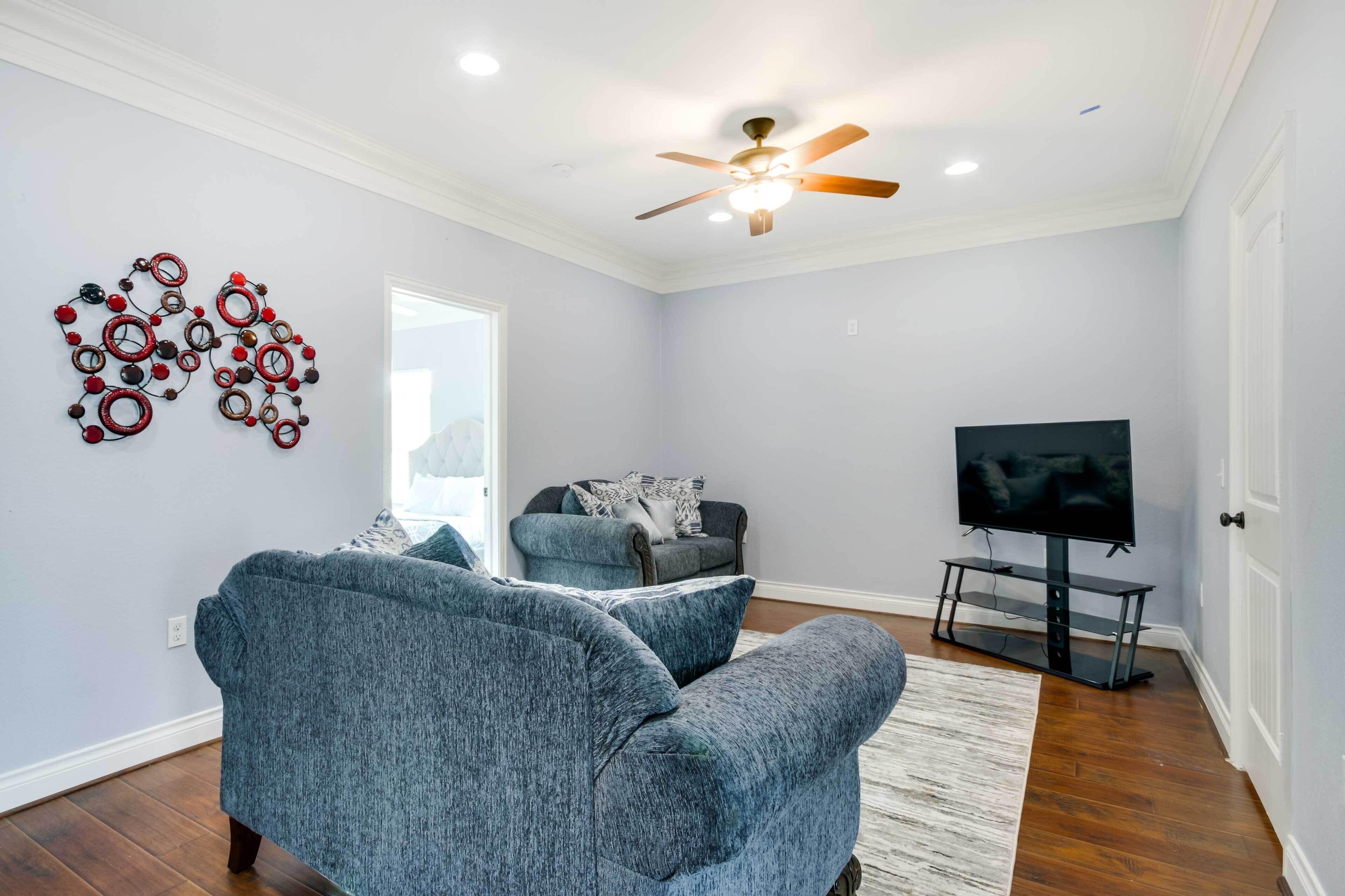 A living room with two blue armchairs, a grey sofa, a ceiling fan, a flat-screen TV on a stand, and a decorative wall piece.