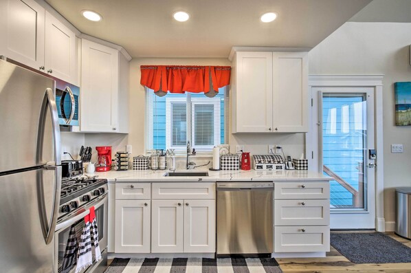 A modern kitchen with white cabinets, stainless steel appliances, a red valance over the window, and a checkered floor mat.