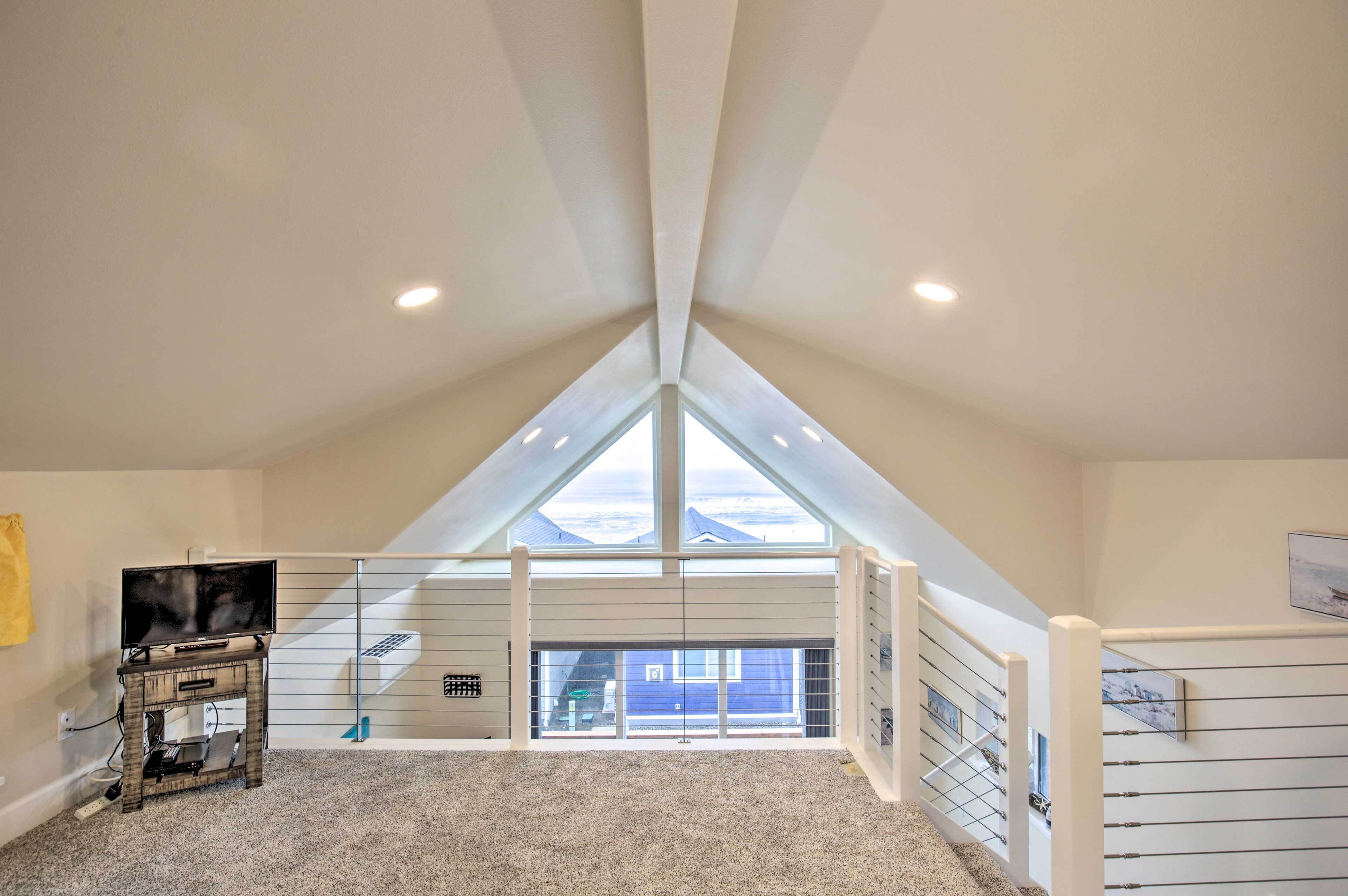A high-ceilinged attic room with a large skylight, a TV on a stand, and a view of a blue house through the window.