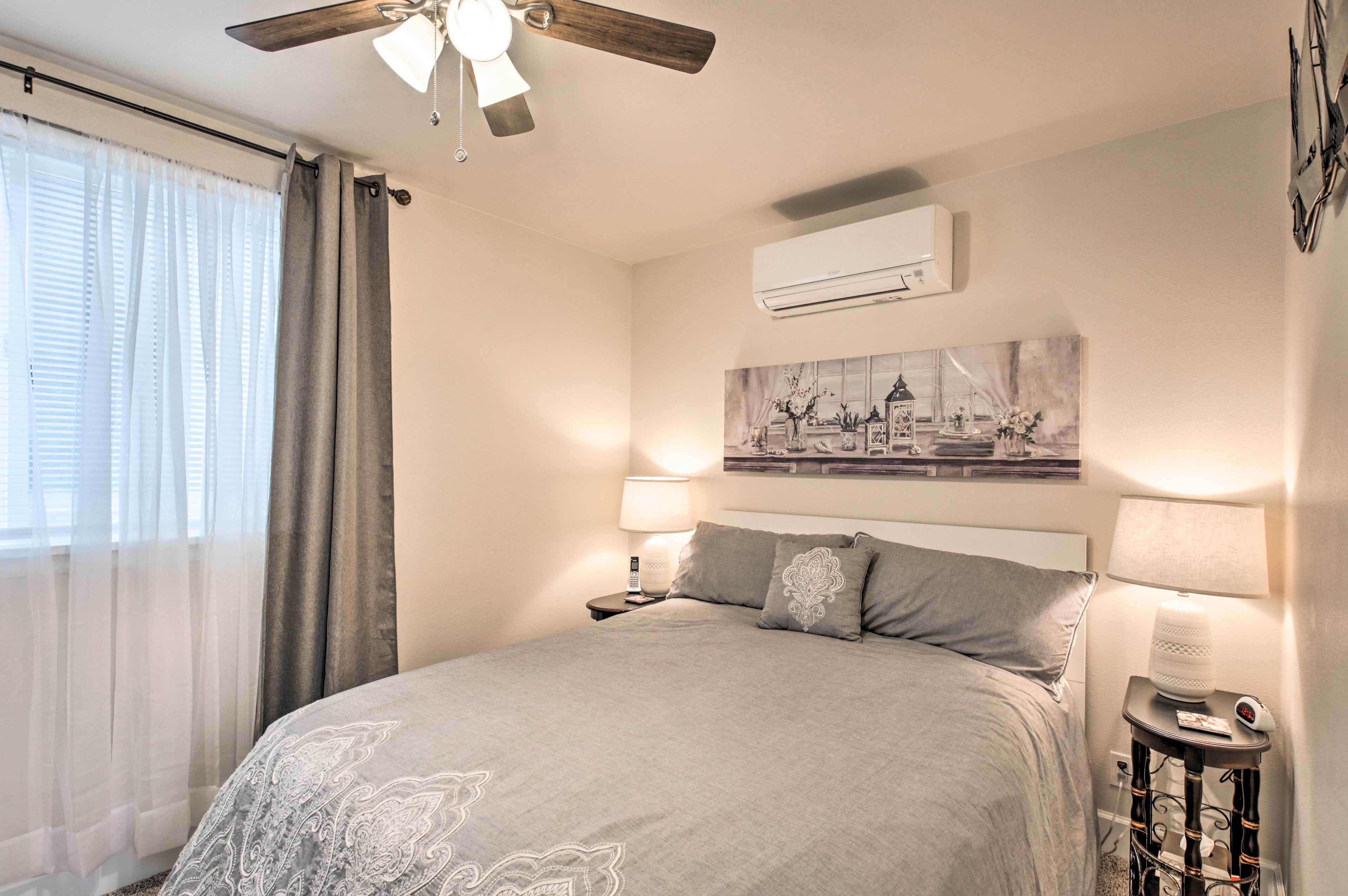 A bedroom with a bed, a ceiling fan, an air conditioning unit, a nightstand with a lamp, and a wall-mounted artwork.