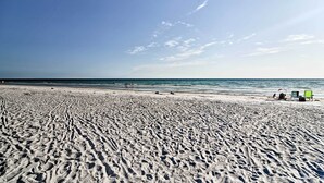 A sandy beach with footprints leading to the water, a few chairs, and a clear sky. - Heated Pool, Steps to Beach: Indian Shores Condo! (Indian Shores)