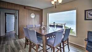 A dining area with a wooden table and chairs, a chandelier, and a large window with a view of the ocean.