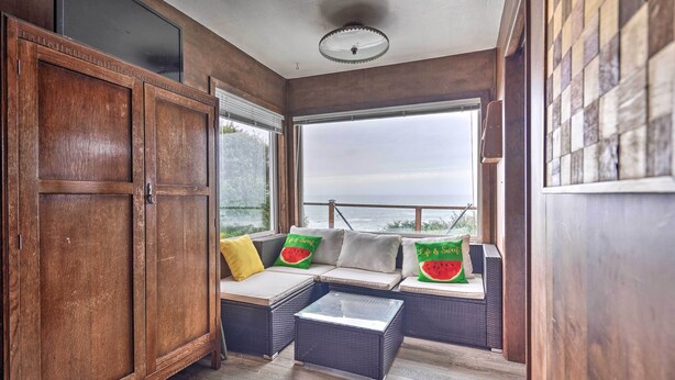 A cozy room with a wooden cabinet, a cushioned bench with pillows, and a view through large windows.