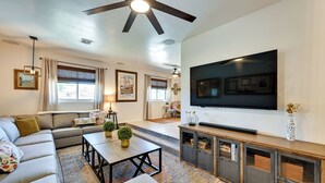 A modern living room with a sectional sofa, a flat-screen TV mounted on the wall, a ceiling fan, and a large window with blinds. - Yuma Vacation Rental w/ Private Pool & Patio! (Yuma)