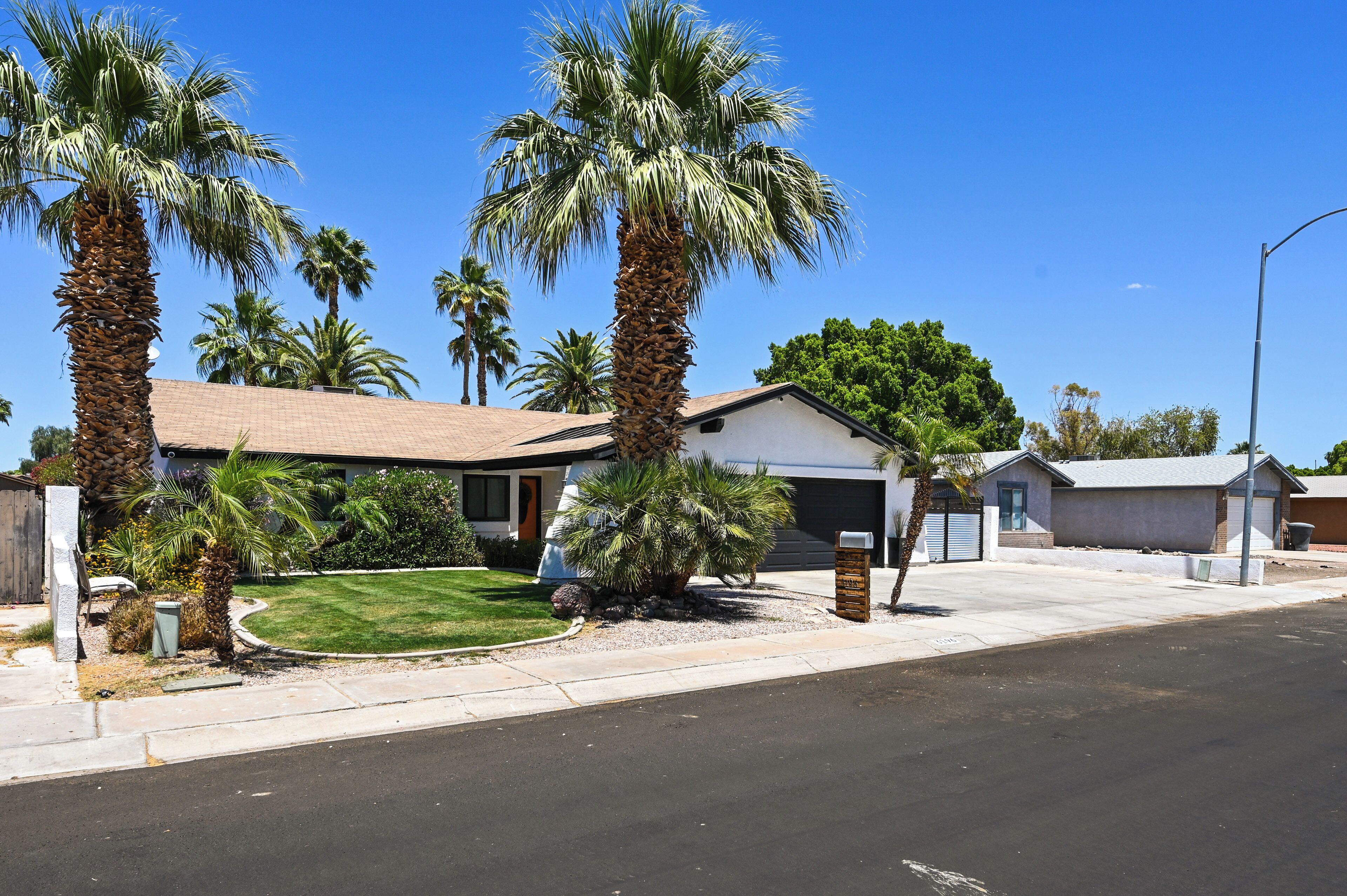 A residential street with palm trees, a house with a garage, and a clear blue sky.