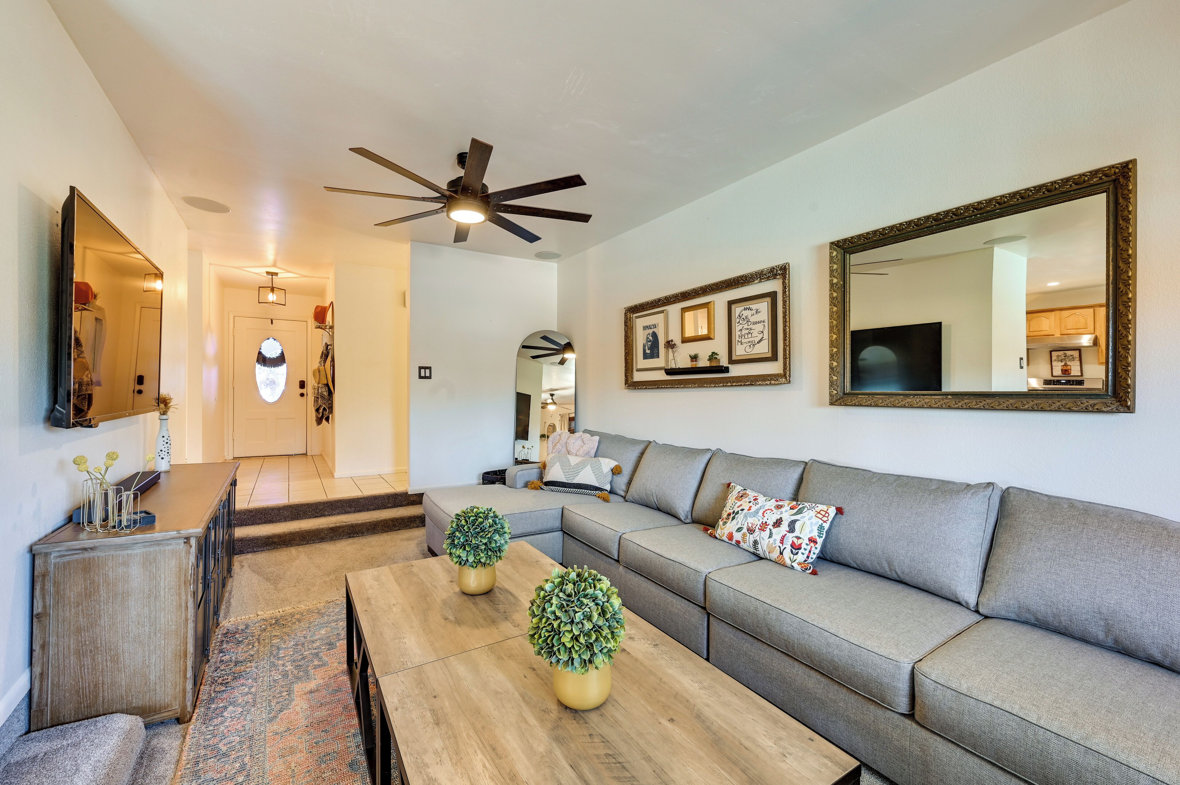 A modern living room with a grey sectional sofa, a wooden coffee table, two potted plants, a ceiling fan, and a large mirror on the wall.