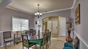 A dining area with a round table, chairs, and a chandelier. There is a window with blinds, a door, and a vase with dried plants.
