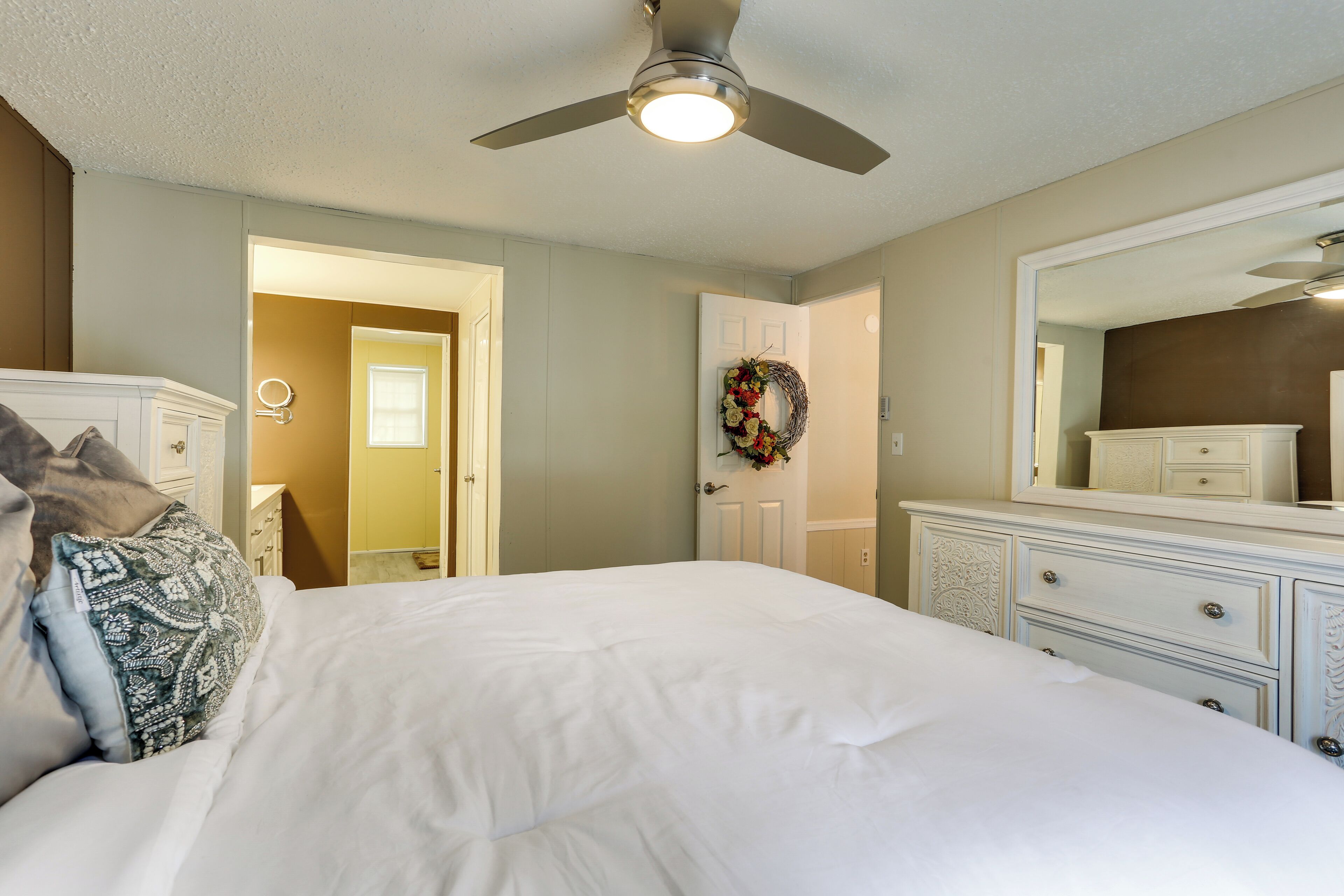 A bedroom with a large bed, a dresser, a mirror, a ceiling fan, and a door leading to another room.