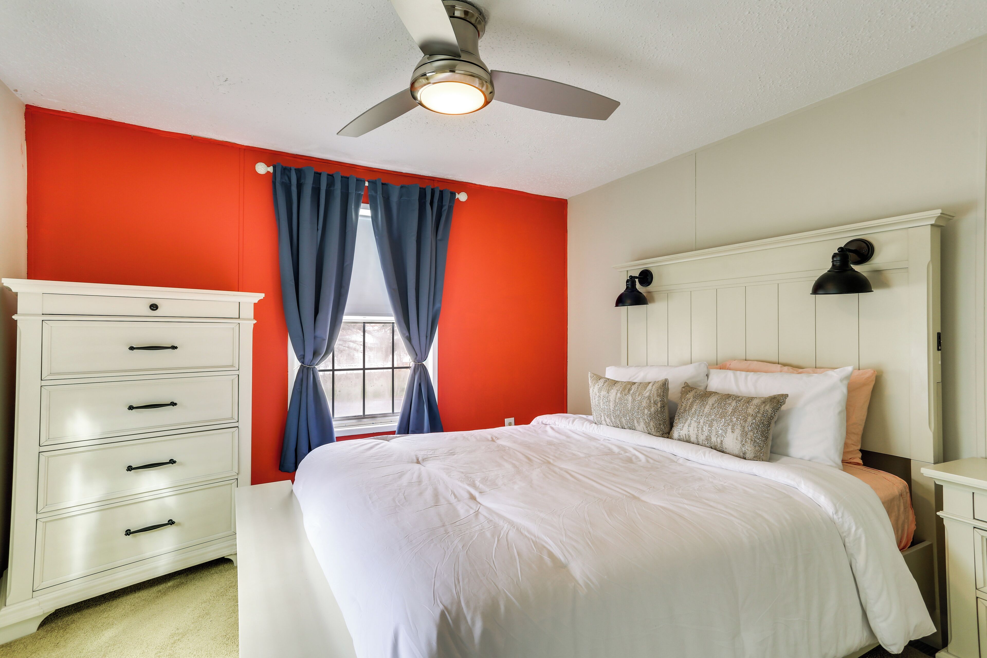 A bedroom with a white bed, a dresser, a ceiling fan, and a window with blue curtains.