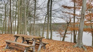 A picnic table in a forested area with a lake in the background. - Walk to Lake! All-season Retreat in Poconos Mtns (Greentown)