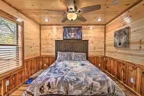 A wooden-paneled room with a bed, a ceiling fan, a mounted TV, and a window with blinds.