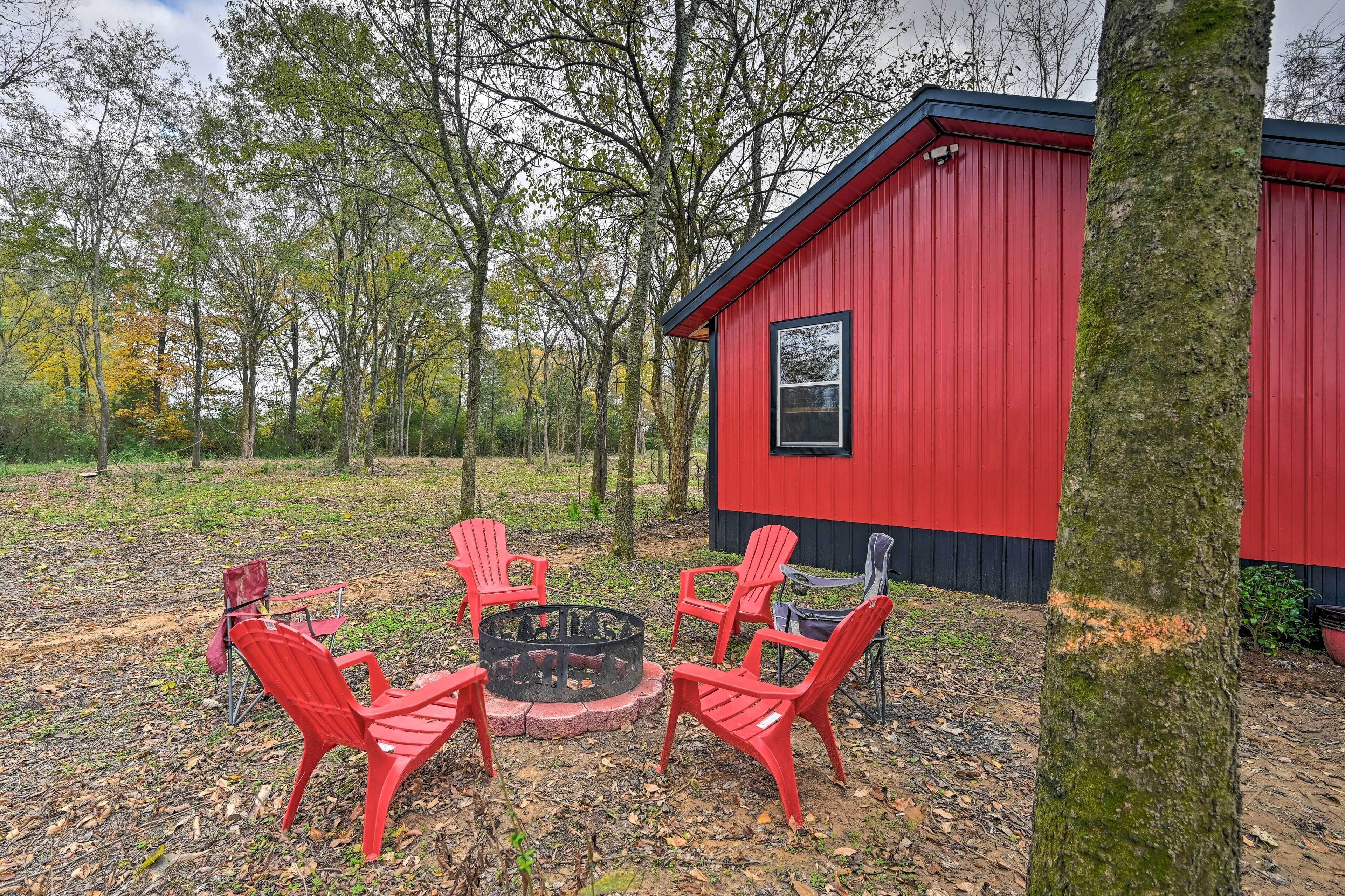 Una cabaña roja con un pozo de fuego y cuatro sillas Adirondack rojas en un área boscosa.